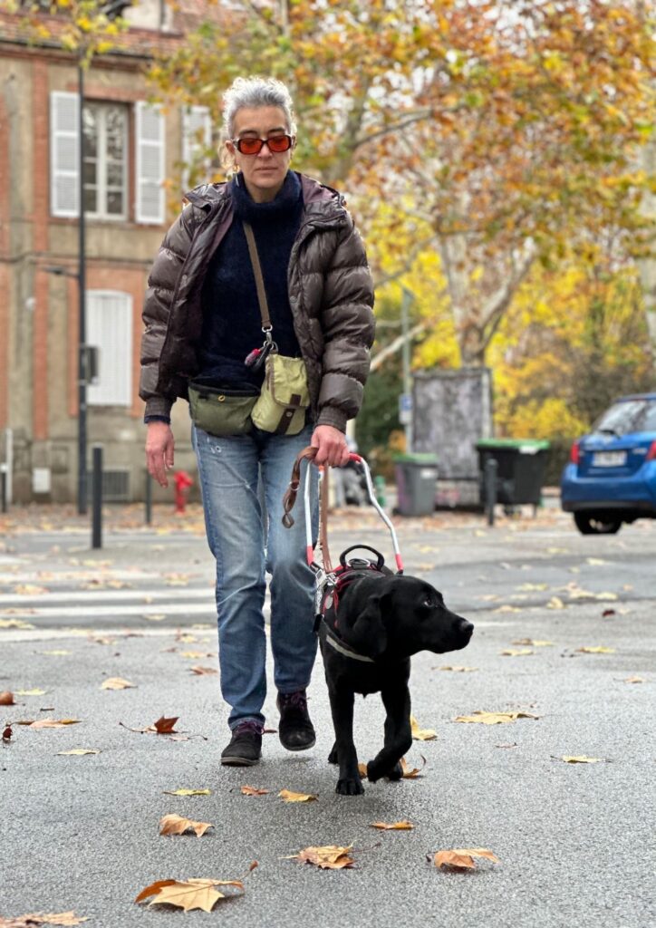 Laurence, maîtresse de chien guide, se promène en ville avec son chien guide Tilt, un labrador noir