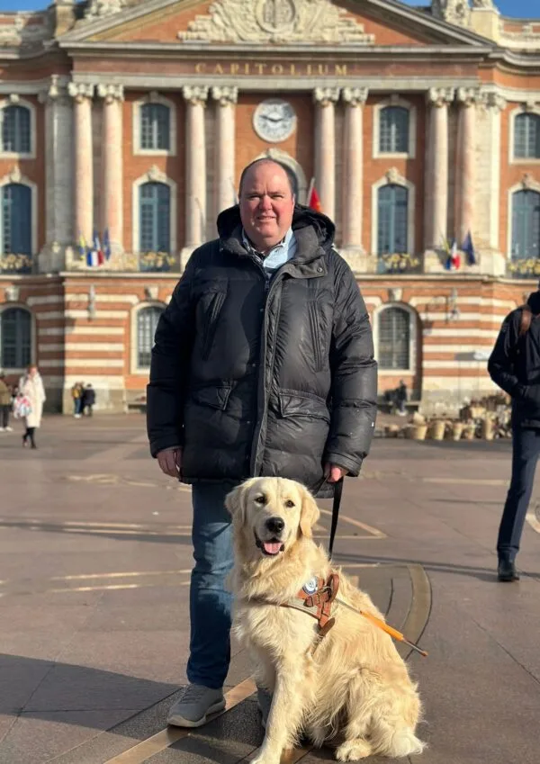 Un homme portant une doudoune noire se tient sur une grande place devant un bâtiment historique en briques rouges et colonnes blanches. À ses côtés, un golden retriever équipé d’un harnais d’assistance est assis calmement face à l’objectif. Le soleil éclaire la scène et quelques personnes apparaissent en arrière-plan