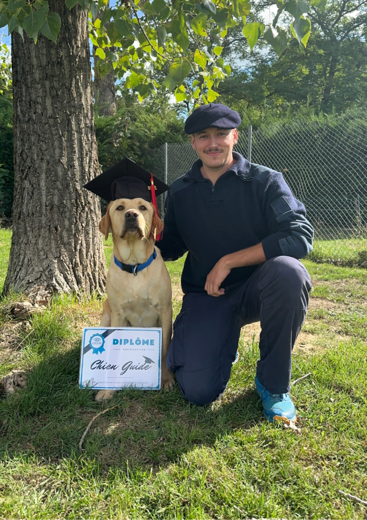 Un homme accroupi à côté d’un chien guide labrador portant une toque de diplômé noire. Le chien est assis devant un panneau indiquant « Diplôme – Chien Guide ». La scène se déroule en extérieur, sous un grand arbre, avec de l’herbe, une clôture et un ciel ensoleillé en arrière-plan.