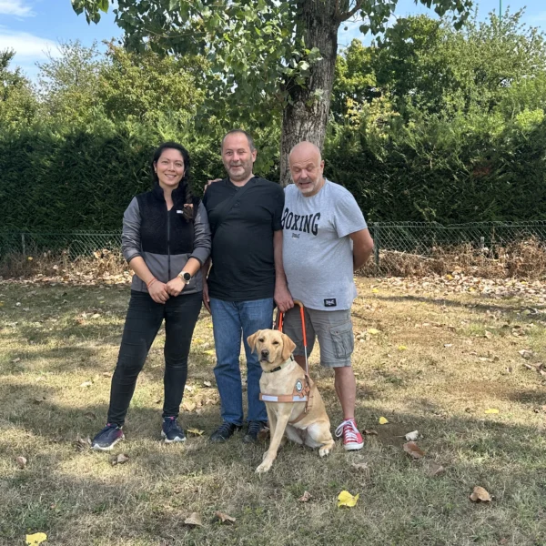 Trois personnes debout dans un jardin ombragé autour d'un labrador sable en harnais de guidage assis, dont un homme tenant une canne blanche.
